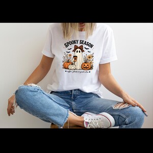May include: A white t-shirt featuring a graphic of a ghost wearing a checkered bow, holding a coffee cup, surrounded by pumpkins, sunflowers, and bats. The text reads SPOOKY SEASON and coffee, ghosts & good vibes in black lettering.