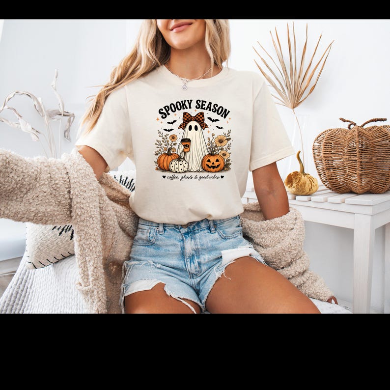 May include: A cream-colored short-sleeve t-shirt featuring a graphic of a ghost wearing a plaid bow, holding a coffee cup, surrounded by pumpkins, sunflowers, and bats. The text reads SPOOKY SEASON and coffee, ghosts & good vibes.