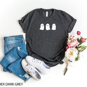 K&ouml;nnte beinhalten: Heather dunkelgraues T-Shirt mit drei wei&szlig;en Geister-Illustrationen. Das Shirt wird auf einem wei&szlig;en Hintergrund mit einer blauen Jeans, wei&szlig;en Turnschuhen und einem Strau&szlig; rosa und wei&szlig;er Blumen gezeigt.