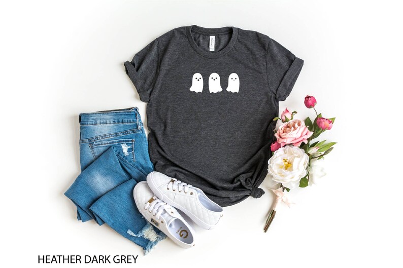 K&ouml;nnte beinhalten: Heather dunkelgraues T-Shirt mit drei wei&szlig;en Geister-Illustrationen. Das Shirt wird auf einem wei&szlig;en Hintergrund mit einer blauen Jeans, wei&szlig;en Turnschuhen und einem Strau&szlig; rosa und wei&szlig;er Blumen gezeigt.