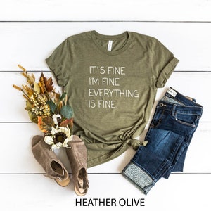 May include: A green t-shirt with the text "It's fine I'm fine Everything is fine." in white lettering. The shirt is on a white wooden background with a pair of brown shoes, a bouquet of dried flowers, and a pair of blue jeans.