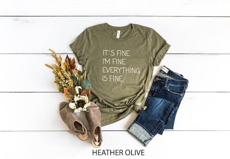 May include: A green t-shirt with the text "It's fine I'm fine Everything is fine." in white lettering. The shirt is on a white wooden background with a pair of brown shoes, a bouquet of dried flowers, and a pair of blue jeans.