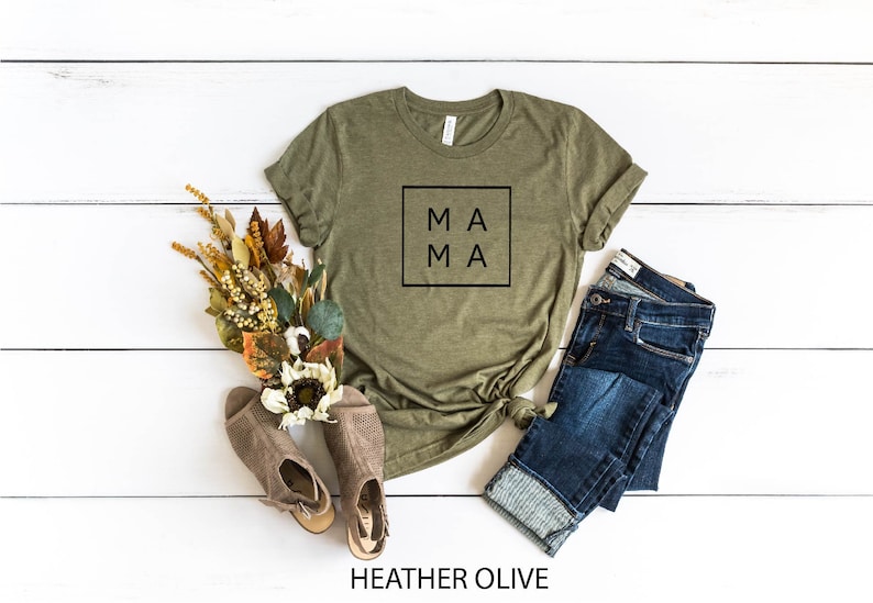 May include: A heather olive t-shirt with the word "MAMA" in a black square on the front. The shirt is laid out on a white wooden surface with a pair of brown sandals, a pair of blue jeans, and a bouquet of dried flowers.