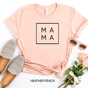 May include: A peach colored t-shirt with the word "MAMA" in a black square on the front. The shirt is laid flat on a white background with a pair of grey shoes, sunglasses, and a bouquet of pink roses.