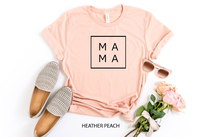 May include: A peach colored t-shirt with the word "MAMA" in a black square on the front. The shirt is laid flat on a white background with a pair of grey shoes, sunglasses, and a bouquet of pink roses.