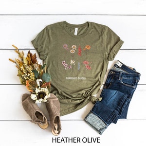 May include: A heather olive t-shirt featuring a floral design and the text "Grandma's Garden". The shirt is styled with blue jeans and brown sandals.