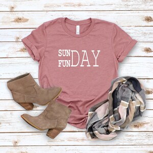 May include: A pink t-shirt with the text "SUN FUN DAY" in white lettering. The shirt is paired with brown ankle boots and a plaid scarf.
