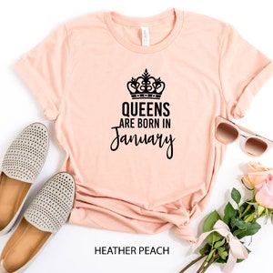 May include: A peach-colored t-shirt with a black crown and the text "Queens are born in January". The shirt is laid flat on a white surface with a pair of beige shoes, sunglasses, and a bouquet of pink roses.