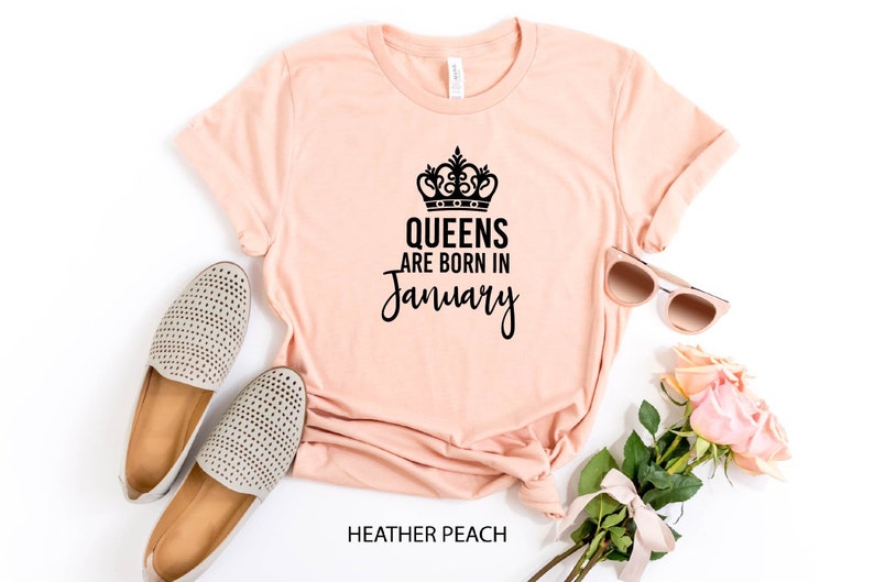 May include: A peach-colored t-shirt with a black crown and the text "Queens are born in January". The shirt is laid flat on a white surface with a pair of beige shoes, sunglasses, and a bouquet of pink roses.