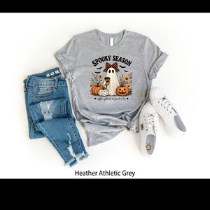 May include: A heather athletic grey t-shirt featuring a graphic of a ghost wearing a plaid bow, holding a coffee cup, surrounded by pumpkins, sunflowers, and bats. The text reads SPOOKY SEASON and coffee, ghosts & good vibes. Blue jeans and white sneakers.