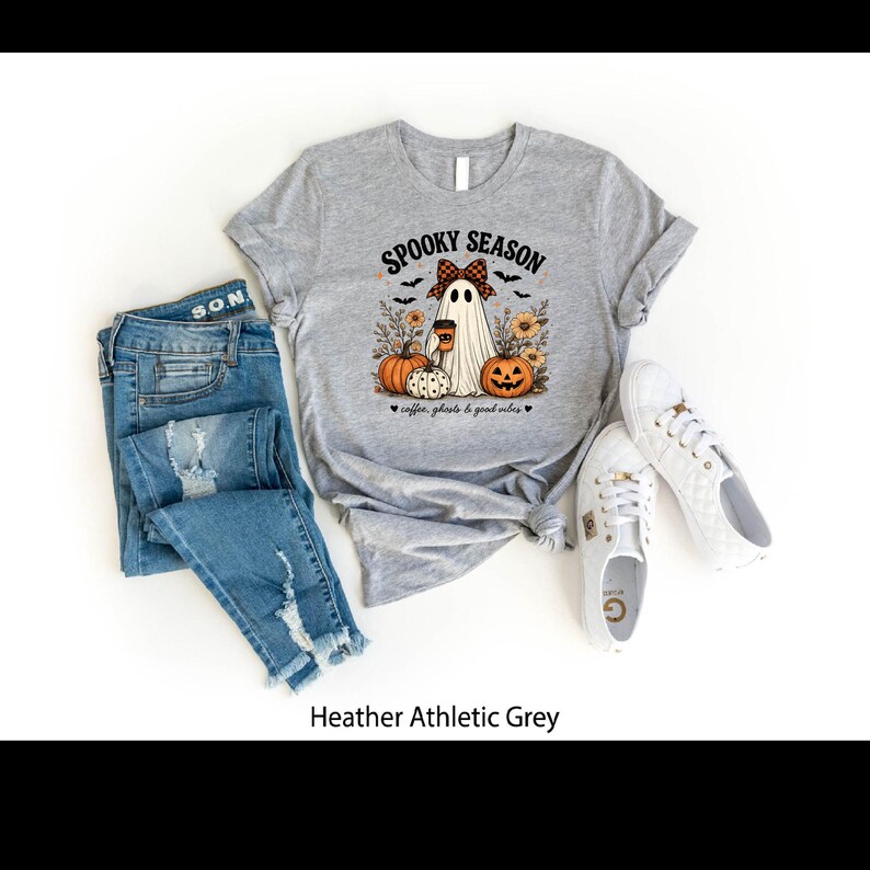 May include: A heather athletic grey t-shirt featuring a graphic of a ghost wearing a plaid bow, holding a coffee cup, surrounded by pumpkins, sunflowers, and bats. The text reads SPOOKY SEASON and coffee, ghosts & good vibes. Blue jeans and white sneakers.