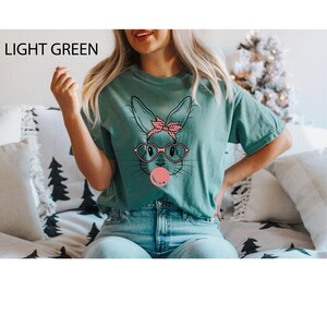 May include: A light green t-shirt with a graphic of a cartoon bunny wearing sunglasses and a pink bow. The bunny is blowing a pink bubble.