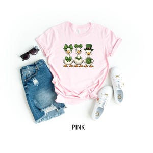 May include: Pink t-shirt featuring three cartoon geese dressed for St. Patrick's Day. The geese wear green bows, shamrock headbands, and a leprechaun hat. The shirt is styled with denim shorts, sunglasses, and white sneakers.