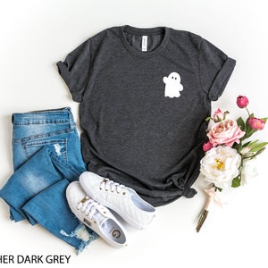 May include: Heather dark grey t-shirt with a white ghost graphic on the front. The shirt is paired with blue jeans and white sneakers.