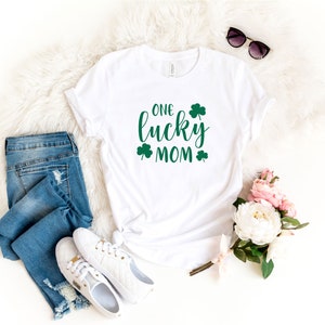 May include: White t-shirt with green shamrock designs and the text "ONE lucky MOM". The shirt is laid out on a white fluffy surface with blue jeans, white sneakers, sunglasses, and pink flowers.