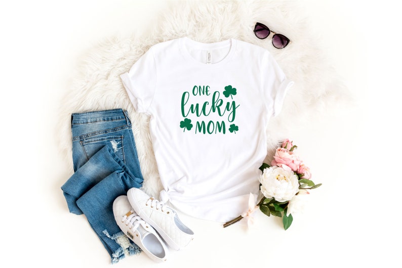 May include: White t-shirt with green shamrock designs and the text "ONE lucky MOM". The shirt is laid out on a white fluffy surface with blue jeans, white sneakers, sunglasses, and pink flowers.