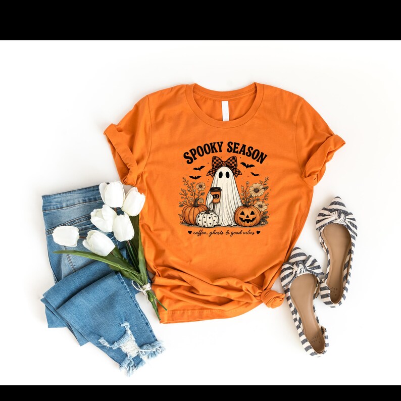 May include: An orange t-shirt featuring a graphic of a ghost wearing a plaid bow, holding a coffee cup, surrounded by pumpkins, flowers, and bats. The text reads SPOOKY SEASON and coffee, ghosts & good vibes. Includes blue jeans and striped flats.