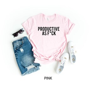 May include: A light pink short-sleeve t-shirt with the black text "PRODUCTIVE AS F*CK" printed on the front, paired with blue distressed denim shorts, white lace-up sneakers, and black sunglasses. The word "PINK" is printed at the bottom.