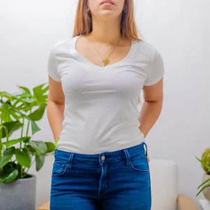 Könnte beinhalten: Ein weißes V-Ausschnitt-T-Shirt mit kurzen Ärmeln und einer goldenen Kettenkette. Die Person trägt eine blaue Jeans.