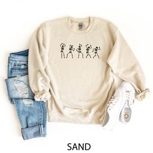 K&ouml;nnte beinhalten: Ein sandfarbenes Sweatshirt mit einem Aufdruck von f&uuml;nf tanzenden Skeletten. Das Sweatshirt wird mit einer blauen Jeans und wei&szlig;en Turnschuhen gezeigt.