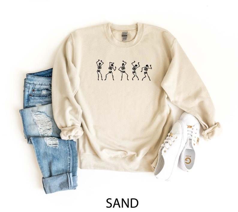K&ouml;nnte beinhalten: Ein sandfarbenes Sweatshirt mit einem Aufdruck von f&uuml;nf tanzenden Skeletten. Das Sweatshirt wird mit einer blauen Jeans und wei&szlig;en Turnschuhen gezeigt.
