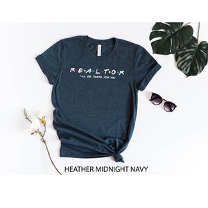 May include: Heather midnight navy t-shirt with the word "REALTOR" in a colorful font, with the phrase "I'll be there for you" underneath. The shirt is styled with a knot at the bottom. Sunglasses and greenery are in the background.