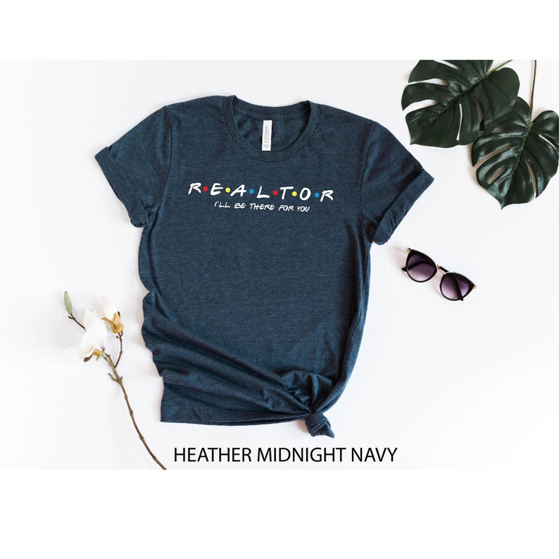 May include: Heather midnight navy t-shirt with the word "REALTOR" in a colorful font, with the phrase "I'll be there for you" underneath. The shirt is styled with a knot at the bottom. Sunglasses and greenery are in the background.
