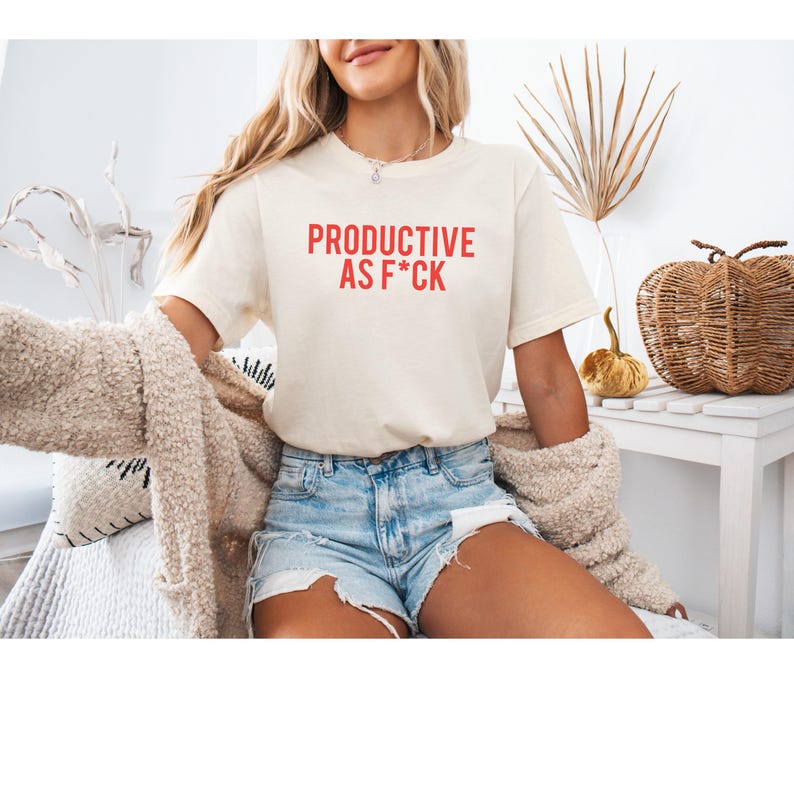 May include: A cream-colored short-sleeve t-shirt featuring the red text "PRODUCTIVE AS F*CK" printed on the chest. The shirt has a crew neckline and a standard fit, paired with light blue denim shorts and a textured beige cardigan sweater.