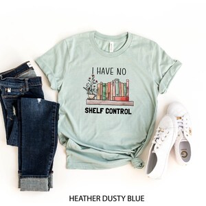 Pu&ograve; includere: Una maglietta blu chiaro con il testo "I have no shelf control" e una grafica di una libreria con libri e una tazza di caff&egrave;. La maglietta &egrave; stesa su uno sfondo bianco con un paio di jeans blu e scarpe da ginnastica bianche.