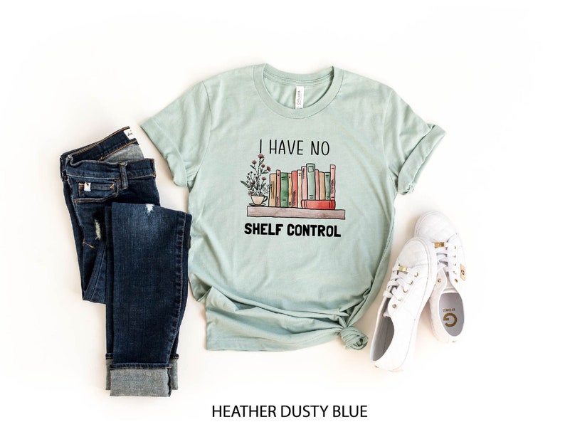 Pu&ograve; includere: Una maglietta blu chiaro con il testo "I have no shelf control" e una grafica di una libreria con libri e una tazza di caff&egrave;. La maglietta &egrave; stesa su uno sfondo bianco con un paio di jeans blu e scarpe da ginnastica bianche.