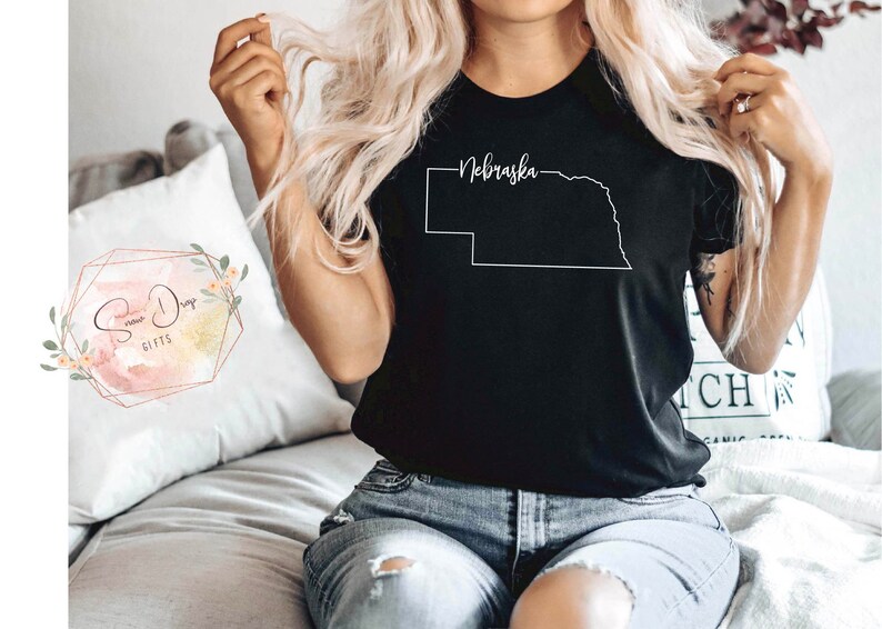 May include: Black t-shirt featuring a white outline of the state of Nebraska with the state name in a cursive font. The shirt is a simple design, perfect for those who love the state. The shirt is a comfortable, casual wear.
