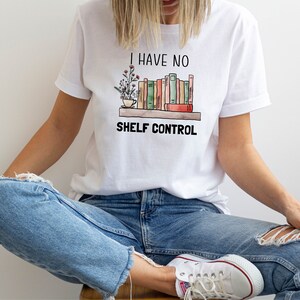 Pu&ograve; includere: Una maglietta bianca con una grafica di una libreria piena di libri e una pianta in vaso. Il testo "I have no shelf control" &egrave; stampato sotto la grafica.