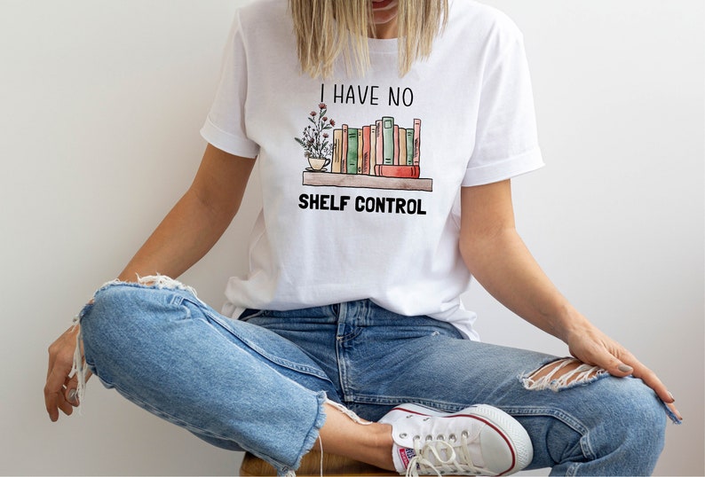 Pu&ograve; includere: Una maglietta bianca con una grafica di una libreria piena di libri e una pianta in vaso. Il testo "I have no shelf control" &egrave; stampato sotto la grafica.