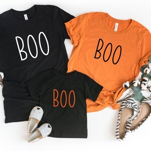 May include: Three t-shirts in black and orange, each with the word "BOO" in a different color. The black shirts have white text, while the orange shirt has black text. The smallest shirt is black with orange text. Halloween-themed apparel.