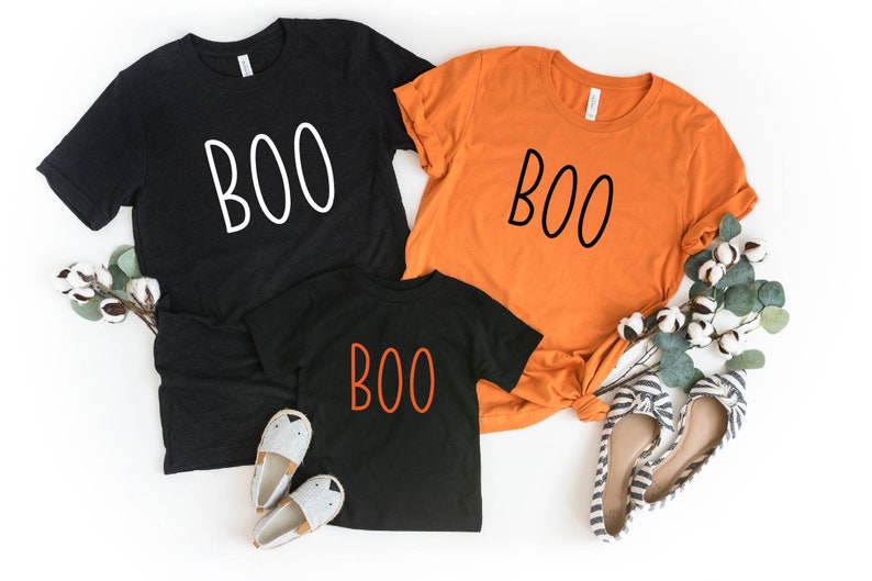 May include: Three t-shirts in black and orange, each with the word "BOO" in a different color. The black shirts have white text, while the orange shirt has black text. The smallest shirt is black with orange text. Halloween-themed apparel.