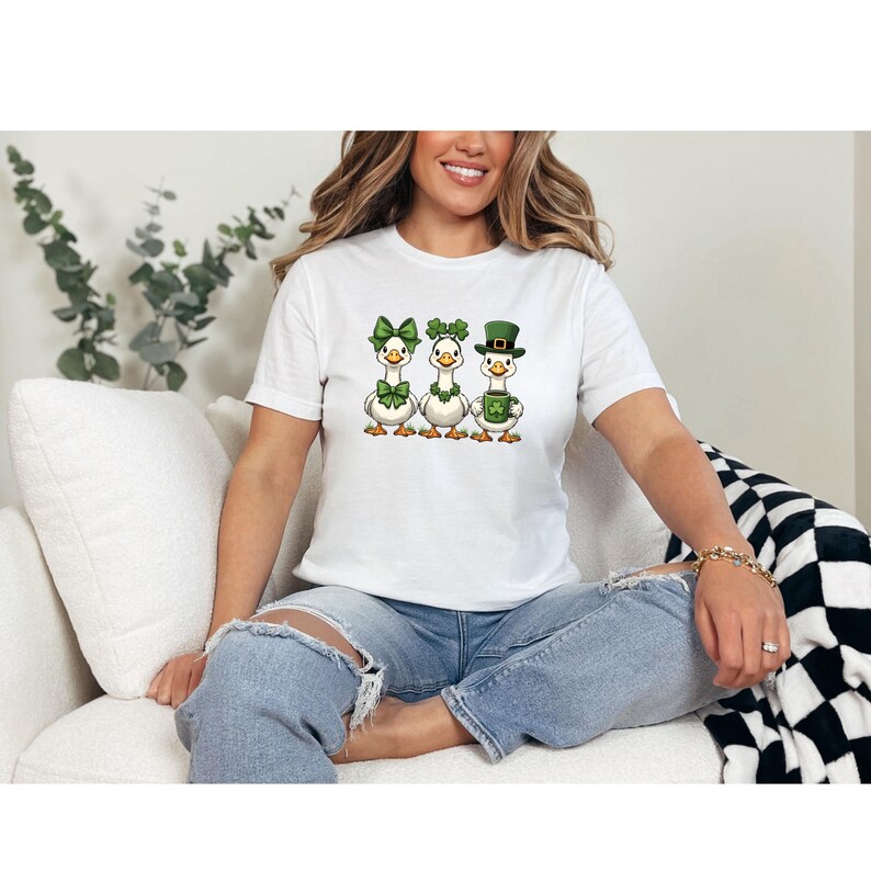 May include: White t-shirt featuring three cartoon geese dressed for St. Patrick's Day. The geese wear green bow ties, shamrocks, and a top hat. The shirt is made of a soft material.