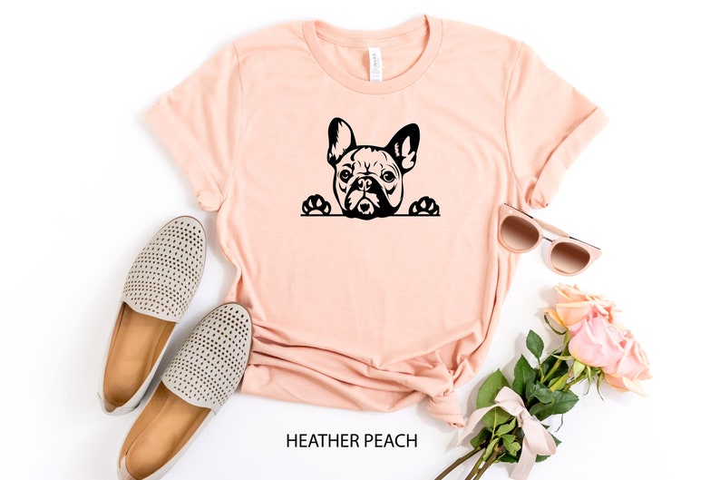 Puede incluir: Una camiseta de color melocot&oacute;n con una silueta negra de un bulldog franc&eacute;s asom&aacute;ndose por la parte superior. El texto "HEATHER PEACH" est&aacute; impreso debajo de la imagen.