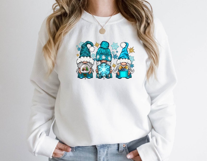 Op de afbeelding: Witte trui met een afbeelding van drie blauwe gnomes die wintermutsen dragen en een sneeuwvlok, een cadeau en een klein huis vasthouden. De gnomes zijn omringd door sneeuwvlokken.