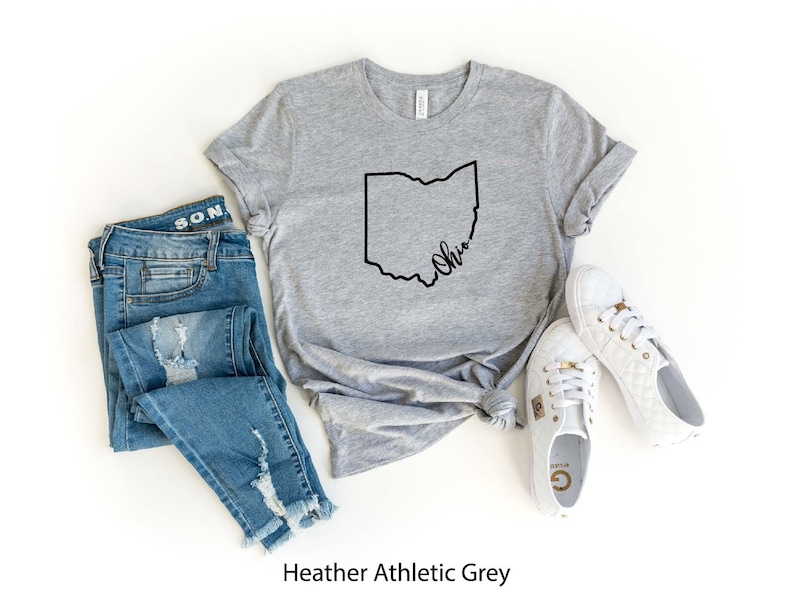 May include: Heather athletic grey t-shirt with a black outline of the state of Ohio and the word "Ohio" written in cursive inside the outline. The shirt is laid flat on a white surface with a pair of distressed blue jeans and white sneakers.