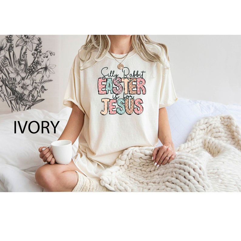 Może przedstawiać: Biała koszulka z napisem "Silly Rabbit Easter is for Jesus" w kolorowych literach z kropkami. Koszulka jest na modelce siedzącej na białym kocu.