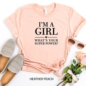 May include: A peach-colored t-shirt with the text "I'm a girl - What's your super power?" in black. The shirt is laid flat on a white background with a pair of beige shoes, sunglasses, and pink roses.