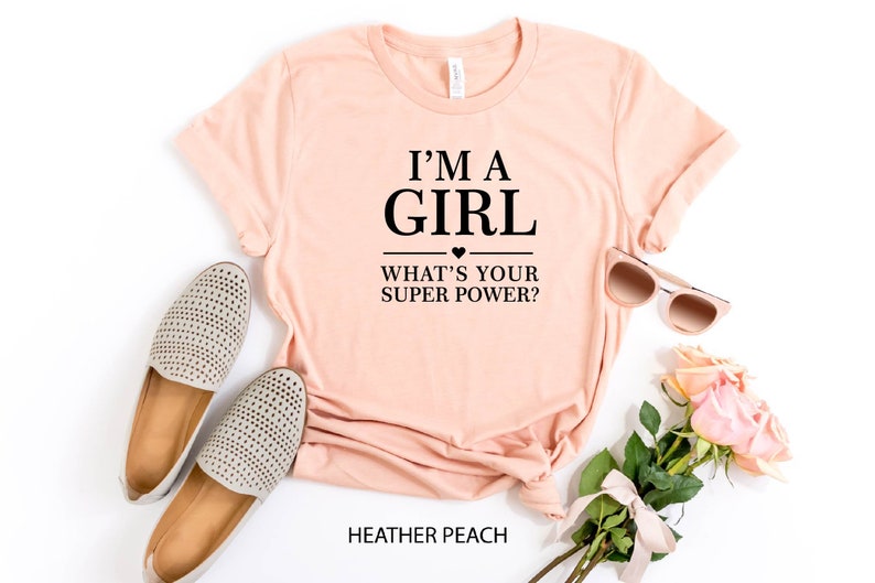 May include: A peach-colored t-shirt with the text "I'm a girl - What's your super power?" in black. The shirt is laid flat on a white background with a pair of beige shoes, sunglasses, and pink roses.