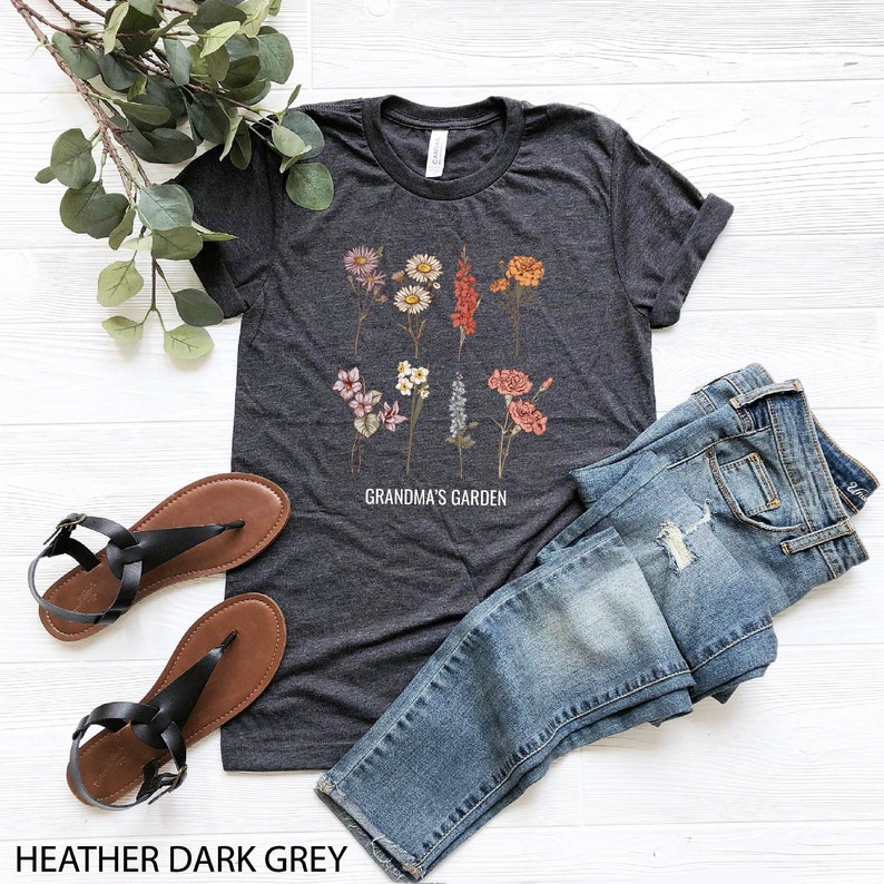 May include: Heather dark grey t-shirt featuring a floral print and the text "Grandma's Garden". The shirt is styled with blue jeans and black sandals.