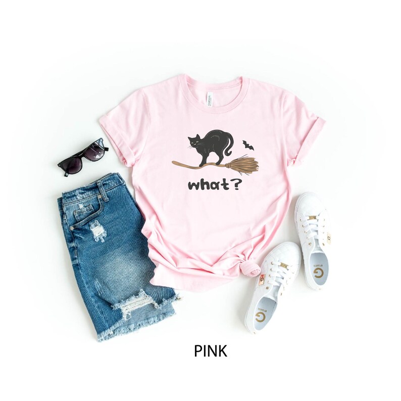 May include: A pink t-shirt with a black cat riding a broom and the word "what?" printed on the front. The image also includes denim shorts, sunglasses, and white sneakers.