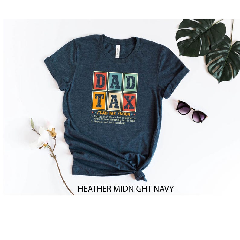 May include: A dark blue t-shirt with a retro-style graphic that says "DAD TAX" and a definition of the term. The text is in a white box with a red, orange, and yellow border.