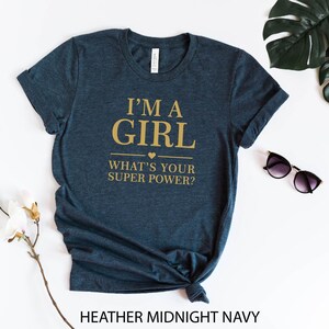 May include: A dark blue t-shirt with the text "I'm a girl what's your super power?" in gold lettering. The shirt is made of a soft, comfortable material and has a relaxed fit.