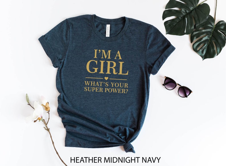 May include: A dark blue t-shirt with the text "I'm a girl what's your super power?" in gold lettering. The shirt is made of a soft, comfortable material and has a relaxed fit.