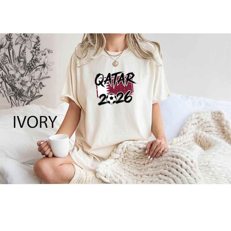 May include: Ivory-colored t-shirt with the text "QATAR 2026" in black, with a soccer ball graphic. The text is set against a maroon and white background. The shirt is short-sleeved.