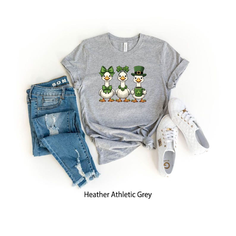May include: Heather athletic grey t-shirt featuring three cartoon geese, each adorned with St. Patrick's Day themed accessories. One goose wears a green bow, another a shamrock, and the last a green hat and mug. Blue jeans and white sneakers complete the ensemble.