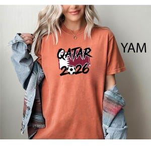 May include: An orange t-shirt with the text "QATAR 2026" in black, with a soccer ball graphic. The shirt features a design incorporating the Qatar flag. The shirt is a short-sleeved, casual style.